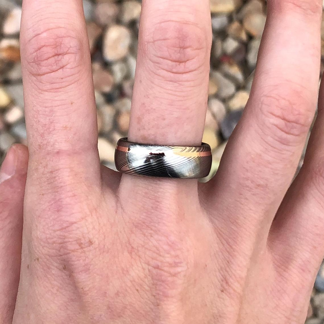 8mm wide Damascus steel ring with an off-centered  rose gold inlay on man&#39;s hand