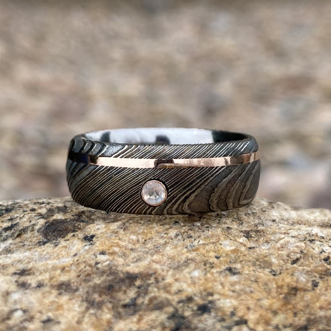 8mm wide black Damascus steel wedding band with an off-centered rose gold inlay with a 3mm round diamond