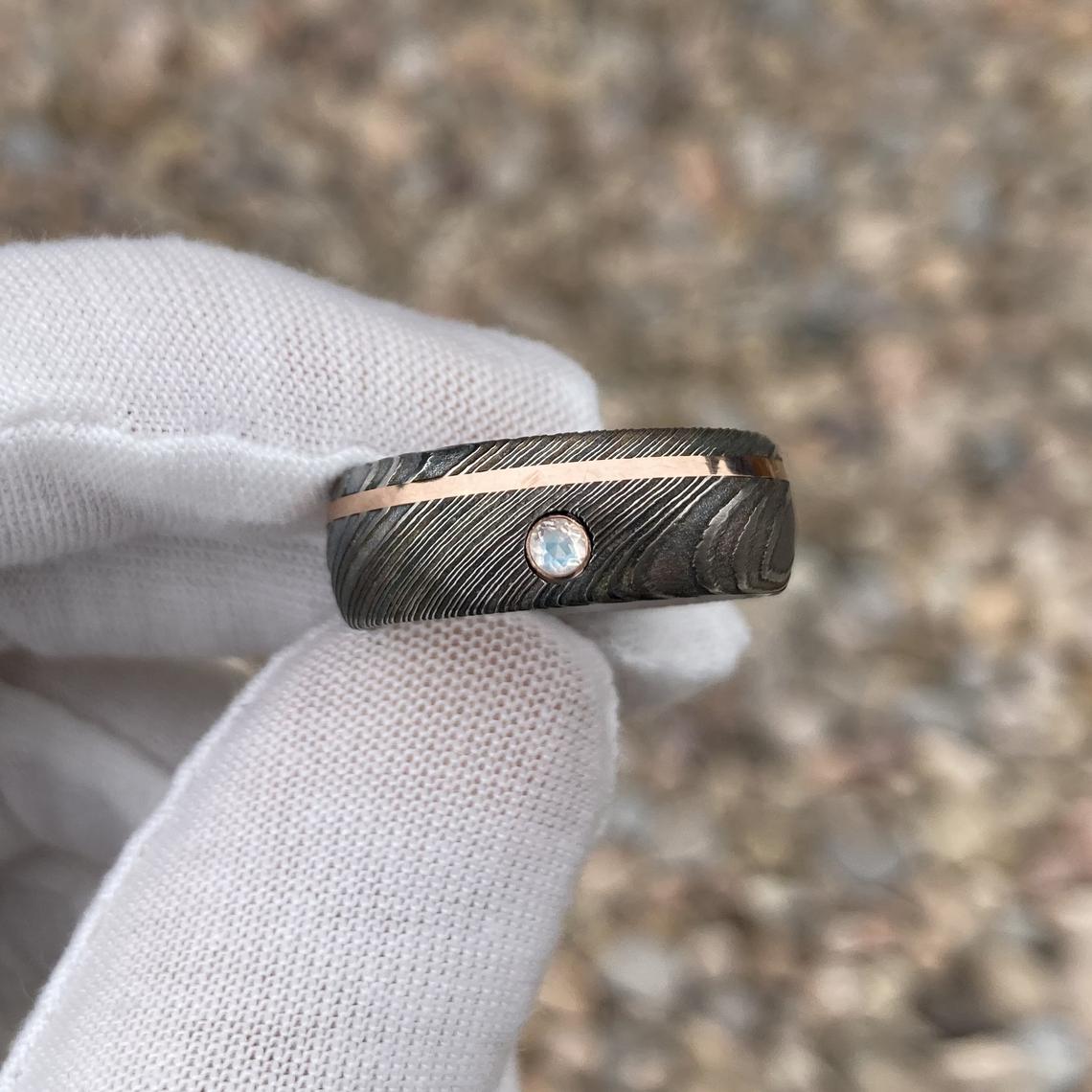 8mm wide black Damascus steel wedding band with an off-centered rose gold inlay with a 3mm round diamond held by white glove