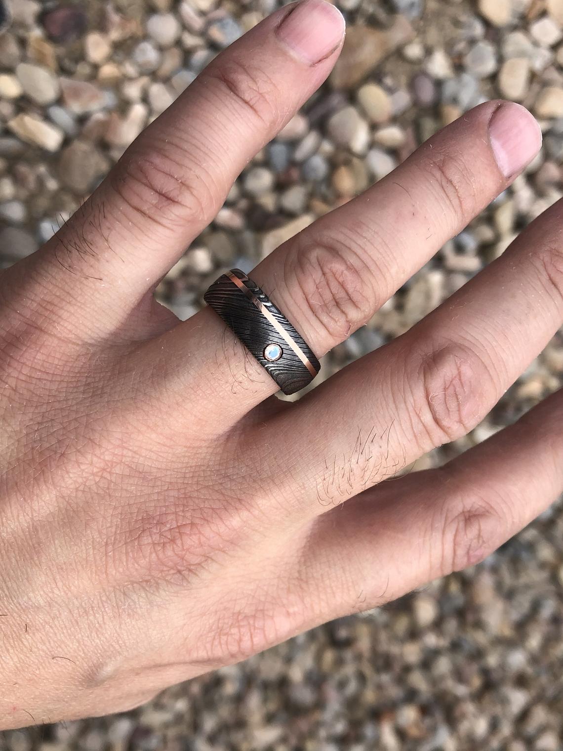 8mm wide black Damascus steel wedding band with an off-centered rose gold inlay with a 3mm round diamond on man&#39;s hand